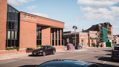 A street scene featuring a brick building with large windows on the left, identified as RBC Financial Group. There are several cars parked along the street, with one prominent black car nearby. The sky is clear with a few clouds. Further down the street, additional buildings are visible with one under renovation covered in green scaffolding. There are a couple of people walking on the sidewalk.