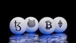 Four spherical objects are placed on a reflective surface with a black background. Each sphere has a cryptocurrency symbol: Tezos, Dogecoin, Bitcoin, and Ethereum. The spheres emit a blue glow, reflecting off the surface below.