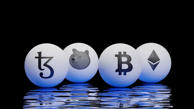 Four spherical objects are placed on a reflective surface with a black background. Each sphere has a cryptocurrency symbol: Tezos, Dogecoin, Bitcoin, and Ethereum. The spheres emit a blue glow, reflecting off the surface below.