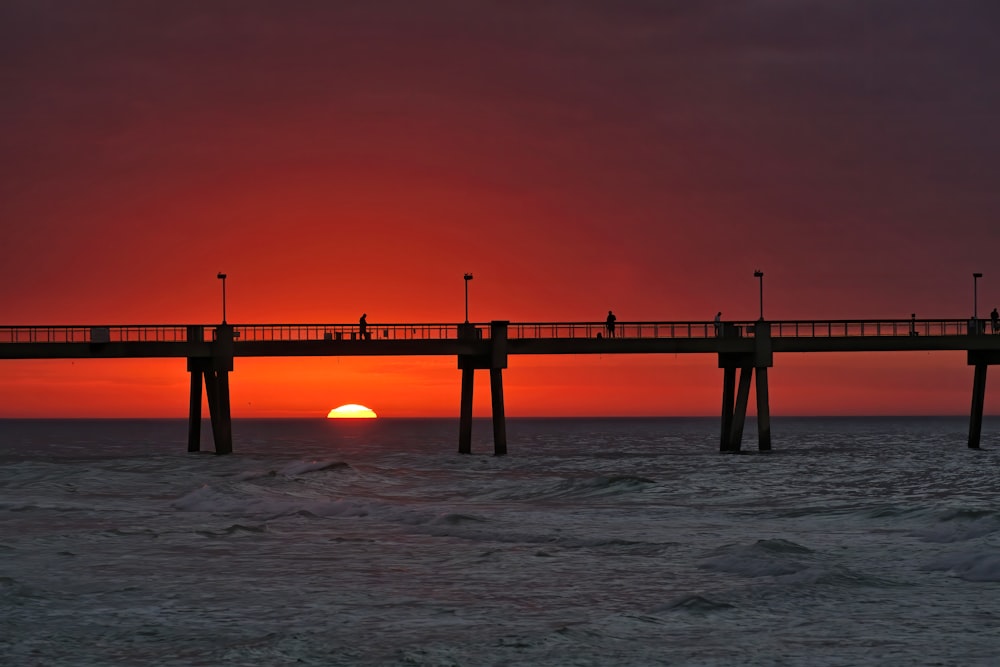 the sun is setting over the ocean with a pier in the foreground