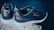 A pair of black and white sneakers are displayed, featuring 'TMC' and a signature in gold on the sides. They rest on a box lined with tissue paper. The shoes have a sleek, modern design with white soles contrasting the dark upper.
