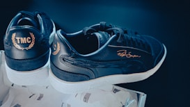 A pair of black and white sneakers are displayed, featuring 'TMC' and a signature in gold on the sides. They rest on a box lined with tissue paper. The shoes have a sleek, modern design with white soles contrasting the dark upper.