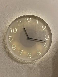 A round, modern-style wall clock with a minimalist design featuring raised, illuminated dots for numbers. The clock has two black hands and a simple glass face, mounted on a light-colored, textured wall.