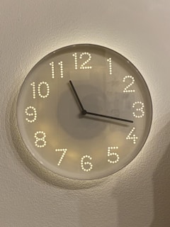A round, modern-style wall clock with a minimalist design featuring raised, illuminated dots for numbers. The clock has two black hands and a simple glass face, mounted on a light-colored, textured wall.