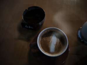 A frothy cappuccino in a brown cup on a wooden table, accompanied by a small bowl filled with dark granules and a sign indicating 64%. The lighting is dim, creating a cozy ambiance.
