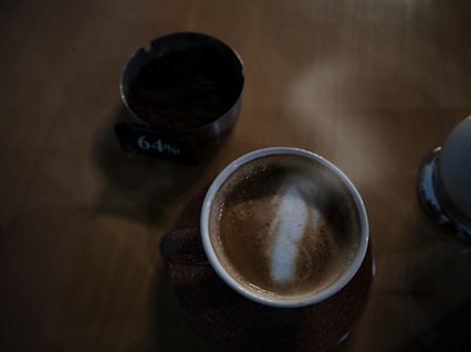 A frothy cappuccino in a brown cup on a wooden table, accompanied by a small bowl filled with dark granules and a sign indicating 64%. The lighting is dim, creating a cozy ambiance.