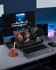 A modern workspace setup with electronic devices including a laptop, desktop monitor, microphone, and a smartphone. The laptop screen displays an editing software with an image of a character holding a shield. The desktop monitor in the background features a dark screen with a masked figure. There is also a microphone on an adjustable boom arm and a small potted plant with a spoon beside the laptop.