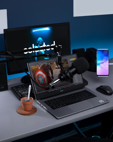A modern workspace setup with electronic devices including a laptop, desktop monitor, microphone, and a smartphone. The laptop screen displays an editing software with an image of a character holding a shield. The desktop monitor in the background features a dark screen with a masked figure. There is also a microphone on an adjustable boom arm and a small potted plant with a spoon beside the laptop.