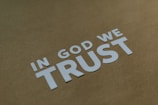 Letters spell out 'IN GOD WE TRUST' in white on a muted brown background, arranged in a diagonal pattern.
