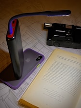 A book is open on a surface with a portable light illuminating the text. A smartphone with a purple case is propped against a black power bank. In the background, a black Panasonic camera is visible.