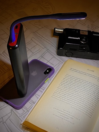 A book is open on a surface with a portable light illuminating the text. A smartphone with a purple case is propped against a black power bank. In the background, a black Panasonic camera is visible.