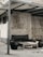 A modern outdoor seating area with a sleek black couch and matching side chairs. The setting includes a simple rectangular coffee table, all arranged under a metal canopy. The background shows a concrete wall, adding an industrial feel to the scene.