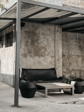 A modern outdoor seating area with a sleek black couch and matching side chairs. The setting includes a simple rectangular coffee table, all arranged under a metal canopy. The background shows a concrete wall, adding an industrial feel to the scene.