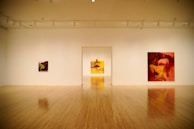 A spacious art gallery room with wooden flooring features three large, colorful paintings with abstract and figurative elements hanging on white walls. The lighting is soft, creating an inviting atmosphere.