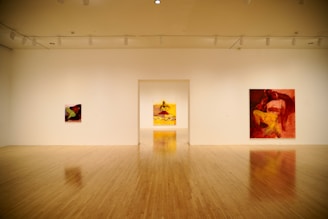 A spacious art gallery room with wooden flooring features three large, colorful paintings with abstract and figurative elements hanging on white walls. The lighting is soft, creating an inviting atmosphere.