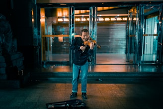 A street musician plays the violin in front of a building entrance during the evening. The scene is dimly lit, with reflections on the polished floor and a few faint lights in the background. The musician's violin case is open on the ground, suggesting he is busking.