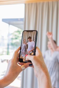 A close-up of a pair of hands holding a smartphone, capturing a photo or video of two people indoors. The background appears slightly blurred, showing a curtain and a bright outdoor area through a window or door. The main focus is on the phone screen displaying the person, one raising a hand.
