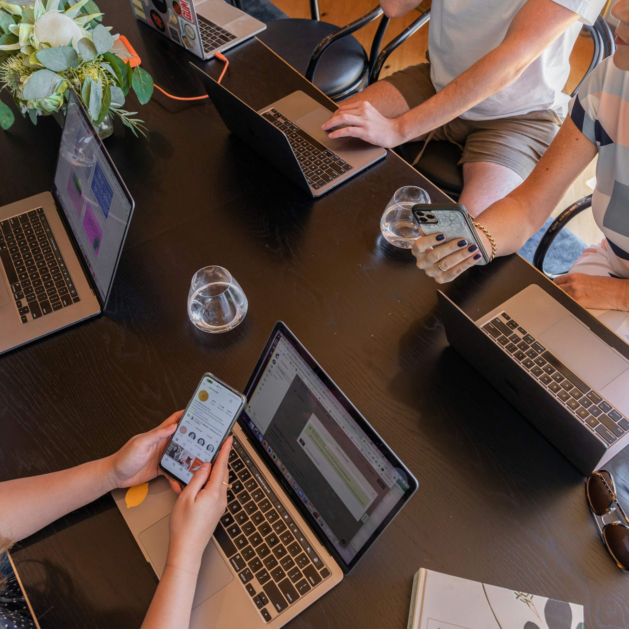Fünf Laptops, zwei Smartphones und drei Menschen an einem Tisch, die zusammenarbeiten.