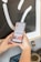Two hands are holding a smartphone displaying a social media profile with follower and following statistics visible. The background includes part of a framed artwork and a decorative object nearby.