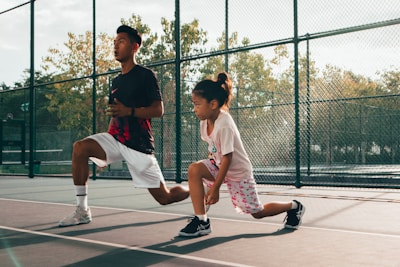 A young child and an adult are performing lunges on an outdoor tennis court. Both are wearing athletic clothing and appear focused. The court is enclosed by a chain-link fence, and trees are visible in the background under clear skies.