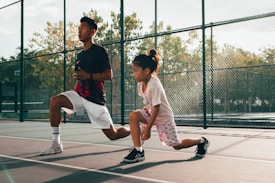 A young child and an adult are performing lunges on an outdoor tennis court. Both are wearing athletic clothing and appear focused. The court is enclosed by a chain-link fence, and trees are visible in the background under clear skies.