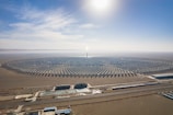 A large, circular solar power plant is situated in a desert landscape. The plant consists of numerous neatly arranged solar panels radiating from a central tower, which reflects sunlight brightly. In the foreground, several buildings and water pools are visible, likely part of the facility's infrastructure. The sky above is clear and blue, with the sun shining intensely.