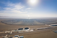 A large, circular solar power plant is situated in a desert landscape. The plant consists of numerous neatly arranged solar panels radiating from a central tower, which reflects sunlight brightly. In the foreground, several buildings and water pools are visible, likely part of the facility's infrastructure. The sky above is clear and blue, with the sun shining intensely.