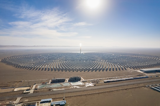 A large, circular solar power plant is situated in a desert landscape. The plant consists of numerous neatly arranged solar panels radiating from a central tower, which reflects sunlight brightly. In the foreground, several buildings and water pools are visible, likely part of the facility's infrastructure. The sky above is clear and blue, with the sun shining intensely.