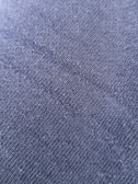 A close-up view of a textured fabric surface with tightly woven fibers forming a diagonal pattern. The material appears to be a dark shade of blue or charcoal grey, with a slightly rough and grainy texture.