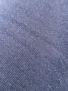 A close-up view of a textured fabric surface with tightly woven fibers forming a diagonal pattern. The material appears to be a dark shade of blue or charcoal grey, with a slightly rough and grainy texture.