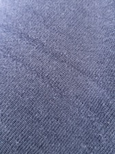 A close-up view of a textured fabric surface with tightly woven fibers forming a diagonal pattern. The material appears to be a dark shade of blue or charcoal grey, with a slightly rough and grainy texture.