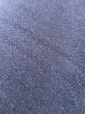 A close-up view of a textured fabric surface with tightly woven fibers forming a diagonal pattern. The material appears to be a dark shade of blue or charcoal grey, with a slightly rough and grainy texture.