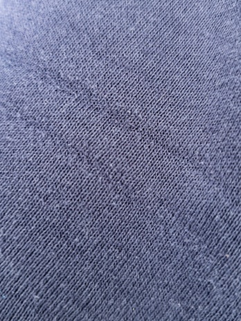 A close-up view of a textured fabric surface with tightly woven fibers forming a diagonal pattern. The material appears to be a dark shade of blue or charcoal grey, with a slightly rough and grainy texture.