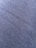 A close-up view of a textured fabric surface with tightly woven fibers forming a diagonal pattern. The material appears to be a dark shade of blue or charcoal grey, with a slightly rough and grainy texture.