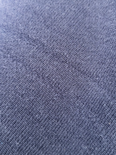 A close-up view of a textured fabric surface with tightly woven fibers forming a diagonal pattern. The material appears to be a dark shade of blue or charcoal grey, with a slightly rough and grainy texture.