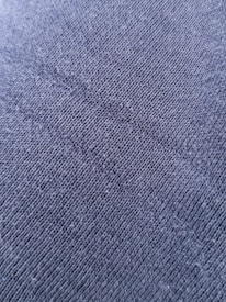 A close-up view of a textured fabric surface with tightly woven fibers forming a diagonal pattern. The material appears to be a dark shade of blue or charcoal grey, with a slightly rough and grainy texture.