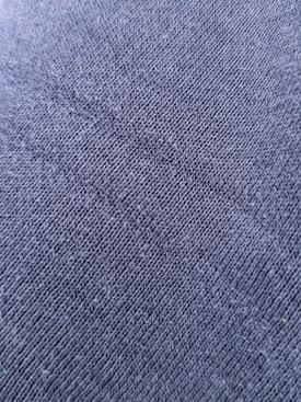 A close-up view of a textured fabric surface with tightly woven fibers forming a diagonal pattern. The material appears to be a dark shade of blue or charcoal grey, with a slightly rough and grainy texture.