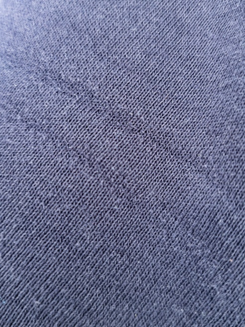 A close-up view of a textured fabric surface with tightly woven fibers forming a diagonal pattern. The material appears to be a dark shade of blue or charcoal grey, with a slightly rough and grainy texture.