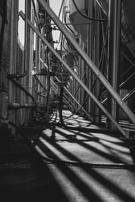 Long metal pipes and beams create a complex industrial structure, casting intricate shadows on the ground. The setting appears to be part of a manufacturing or processing plant with visible cables and conduits.