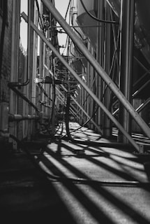 Long metal pipes and beams create a complex industrial structure, casting intricate shadows on the ground. The setting appears to be part of a manufacturing or processing plant with visible cables and conduits.