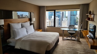 A modern hotel room with a large bed adorned with white pillows and sheets. A sleek glass desk with a chair is situated near a large window that offers a view of a cityscape with tall buildings. The room features a minimalist design with neutral colors and contemporary furniture, including a wall-mounted television and decorative items on a small shelf.