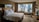 A modern hotel room with a large bed adorned with white pillows and sheets. A sleek glass desk with a chair is situated near a large window that offers a view of a cityscape with tall buildings. The room features a minimalist design with neutral colors and contemporary furniture, including a wall-mounted television and decorative items on a small shelf.