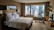 A modern hotel room with a large bed adorned with white pillows and sheets. A sleek glass desk with a chair is situated near a large window that offers a view of a cityscape with tall buildings. The room features a minimalist design with neutral colors and contemporary furniture, including a wall-mounted television and decorative items on a small shelf.