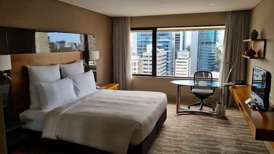 A modern hotel room with a large bed adorned with white pillows and sheets. A sleek glass desk with a chair is situated near a large window that offers a view of a cityscape with tall buildings. The room features a minimalist design with neutral colors and contemporary furniture, including a wall-mounted television and decorative items on a small shelf.