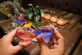 Two hands hold small, colorful glasses filled with liquid, possibly sake, on a wooden table. Behind them are traditional Japanese dishes with ornate presentation, including raw fish served on wooden plates. Greenery decorates the setting, with bottles in the background adding to the atmosphere.