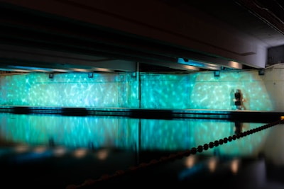 A dimly lit indoor space features a wall illuminated with vibrant turquoise and white light patterns. A pool or body of water reflects the lights, creating a theatrical atmosphere. There is a person visible, standing near the lit wall, adding a sense of scale to the scene.