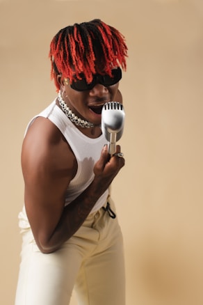 A person with red and black braided hair is enthusiastically singing into a retro-style microphone. They are wearing sunglasses, a white sleeveless top, and cream-colored pants. The background is a soft beige color, accentuating their dynamic pose and energetic expression.