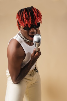 A person with red and black braided hair is enthusiastically singing into a retro-style microphone. They are wearing sunglasses, a white sleeveless top, and cream-colored pants. The background is a soft beige color, accentuating their dynamic pose and energetic expression.