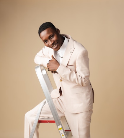 A person wearing a light-colored suit is smiling and leaning on a metal stepladder against a beige background. The individual appears happy and confident.