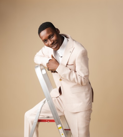 A person wearing a light-colored suit is smiling and leaning on a metal stepladder against a beige background. The individual appears happy and confident.